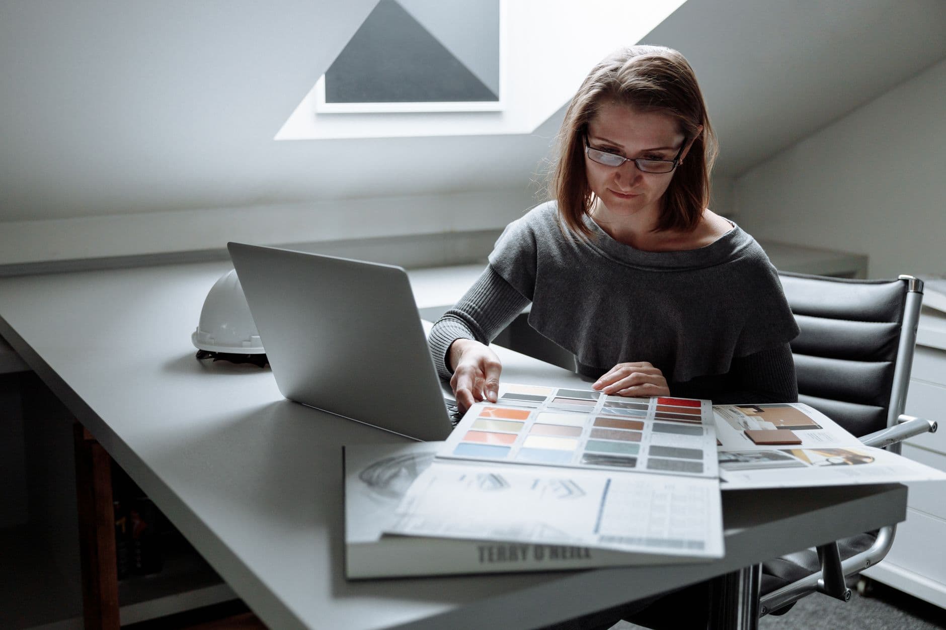 A woman at a laptop working through a sheet of paint color samples, a white hard hat on the desk beside her, the kind of selection this template tracks until it is ordered.